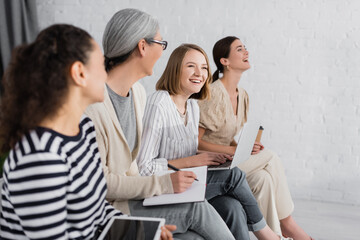 multicultural businesswomen smiling while holding gadgets during seminar