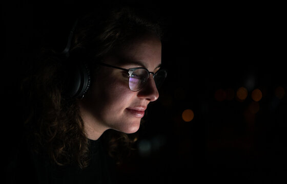 Woman with glasses and headphones surfing internet in night. Screen reflected.