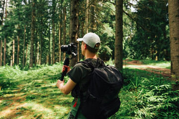 Young male hiker in casual clothes stands in a mountain forest with a camera on a stabilizer and makes a video. Video operator captures content on camera during th