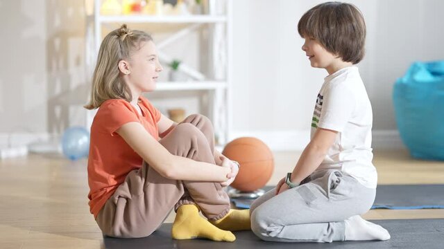 Side view of positive laughing Caucasian boy and girl sitting on exercise mat face to face talking. Portrait of happy carefree brother and sister or friends chatting on weekends at home after training