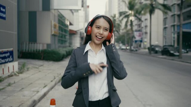  Asian woman, businesswoman, beautiful eyes, white skin, black suit, red wireless headphones. On mobile, smartphone Walking listening to music with pleasure On the side of the road in the city

