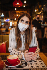 Beautiful young adult woman with protective face mask sitting in a nice restaurant and using her smart phone. She is alone and serious. Coronavirus pandemic and social distancing problems.