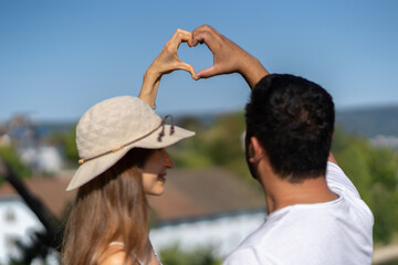 Couple in love raising arm up making heart together in the blue sky. Landscape in background.