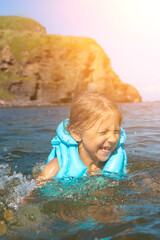 Little girl in a life jacket on a bright summer day bathes in the sea. Child splashing in the water.