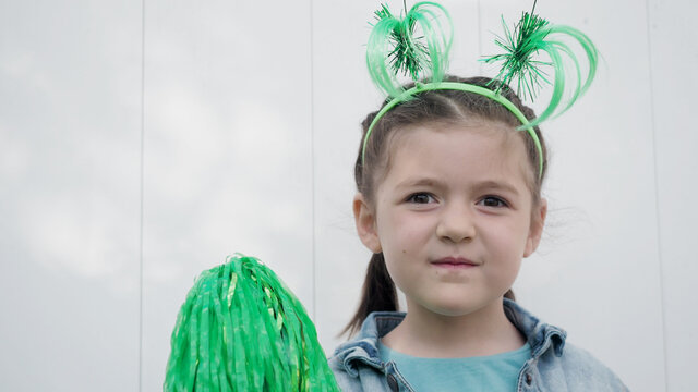 Cute happy brown hair child with green funny horns playing with green pom poms white wall background, celebrating saint patrick's day.