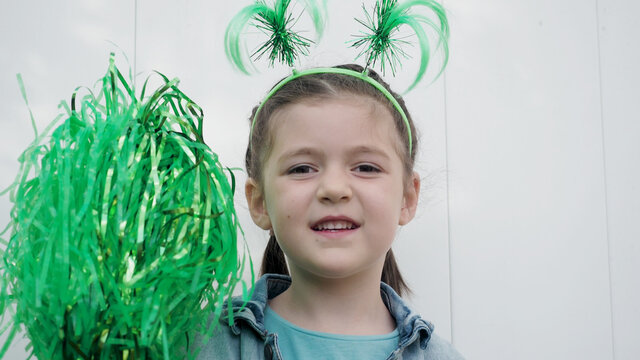 Cute happy brown hair child with green funny horns playing with green pom poms white wall background, celebrating saint patrick's day.