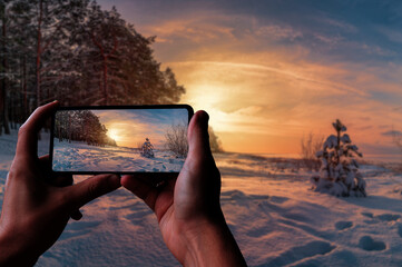 Tourist taking photo of sunset over winter landscape with covered in snow pine and fir trees against dramatic evening light. Snowy Baltic sea coast.