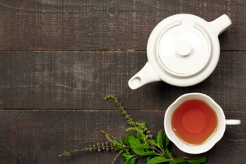 Tulsi or holy basil tea in white cup and teapot with fresh tulsi branch on wooden background top view. Ayurvedic medicine in India.