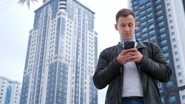 Handsome man in leather jacket with phone on background of tall houses.