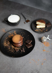 Round chocolate brownie on a black plate and cocoa powder on a gray concrete table. Sweets and coffee background.