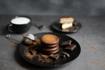 Round chocolate brownie on a black plate and cocoa powder on a gray concrete table. Sweets and coffee background.