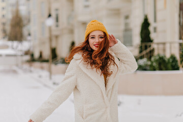 Playful ginger girl in knitted hat looking to camera. Outdoor portrait of young woman posing in winter.