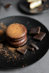 Round chocolate brownie on a black plate and cocoa powder on a gray concrete table. Sweets and coffee background.