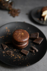 Round chocolate brownie on a black plate and cocoa powder on a gray concrete table. Sweets and coffee background.