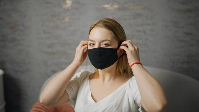 Beautiful young woman with blue eyes putting on protective black mask and looking at you