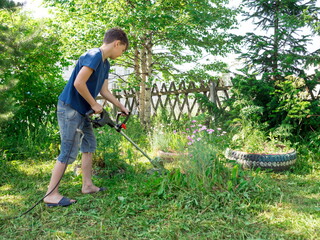A boy, 11 years old, mows the lawn with an electric scythe near the trees in the yard of a house on a sunny summer day.