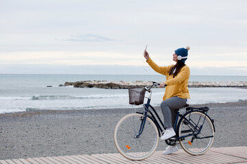 Elegant woman riding a bicycle and using a cell phone. By the sea, yellow sweater and headphones. space for text
