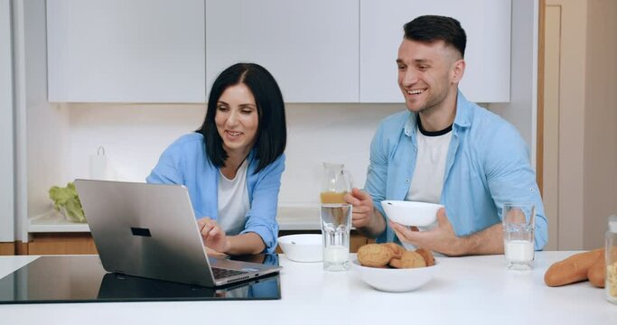 Good-looking happy smiling married couple using computer to revision funny videos while have breakfast in contemporary kitchen