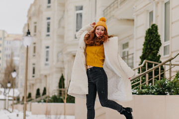 Funny girl in yellow hat dancing in cold day. Excited ginger woman jumping during winter photoshoot.