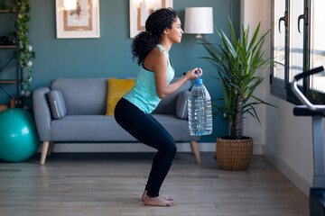 Afro beautiful gymnast woman doing exercices lifting eight liters water bottles in living room at home.