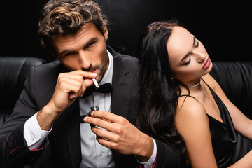 elegant man lightning cigarette and looking at camera near seductive woman sitting with closed eyes isolated on black