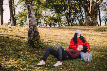 Portrait sweet couple of teenage sitting on hill, asian man with maroon sweater lay down on lap  young woman with red sweater. Romantic mood and sweet emotional relationship in summer with copy space