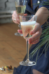 Woman sits on wooden floor, holds glass of champagne, and serves the second glass to viewer. Celebration in informal setting. Concept of romantic evening, wedding anniversary, bachelorette party.