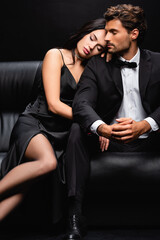 sensual woman with closed eyes leaning on shoulder of elegant man sitting on leather couch isolated on black