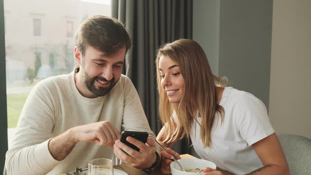 Focused man and woman are using smartphone while eating breakfast sitting at the table in the kitchen at home in the morning