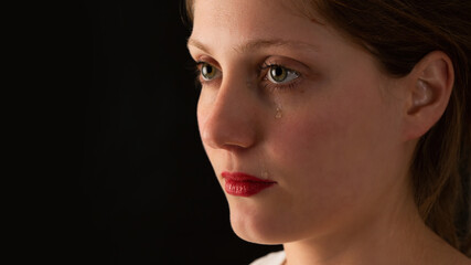 Fototapeta premium Sad woman crying on black background from close up. Unhappy girl with tears falling from eye on dark backdrop. Younge female feeling upset.