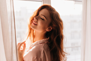 Indoor photo of sensual red-haired girl smiling with eyes closed. Pretty cherful woman posing near window.