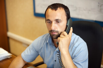 A young employee sits in a chair at a desk in the office and talks on a wired phone. This is a man...