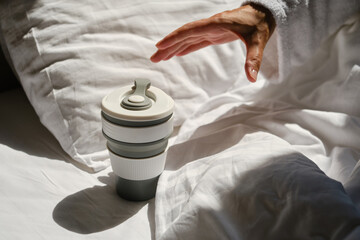 The girl reaches with her hand for a cup of morning coffee. Eco cup for hot drink. Happy lazy girl wakes up in a cozy white bed. Relaxation in the bedroom. Sunlight on linens. Pillow and blanket
