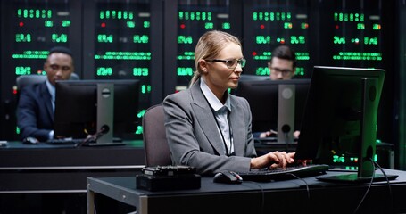 Smart female IT worker with glasses sitting in server room behind workplace, working with database on computer. System software concept.