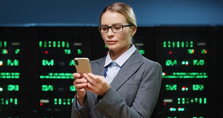 Beautiful female IT programmer with glasses stands in server room holding smartphone and intently...
