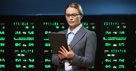 Smart female IT worker in glasses standing in the middle of server room holding tablet and enters data into database.