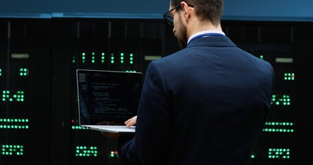 Smart IT worker in suit stands in office holding laptop and inspects technical condition of servers...