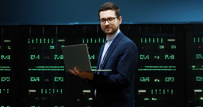 Portrait of IT worker with glasses standing in server room holding laptop and looking at camera smiling. Concept of system support.