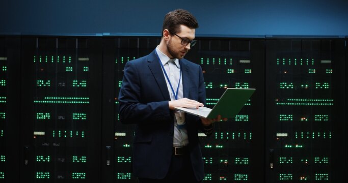 System administrator in glasses and suit standing in the middle of server room holding laptop and enters data into database inspecting technical characteristics of device.