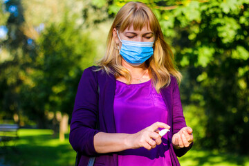 Mature middle-aged woman in blue medical mask, violet blouse washes hands with a gel antiseptic spray isolated over nature park background. web banner, copy space. Overall plan