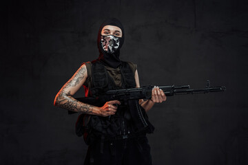 Tattooed and seductive woman in black armour and weared with mask poses in dark background holding assault rifle.