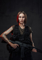 Beautiful woman with short haircut dressed in black armour with dark shirt holding assault rifle in dark background.