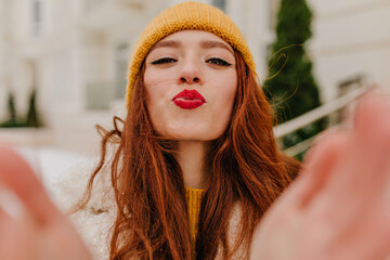 Ecstatic european girl in yellow hat posing in winter. Elegant ginger female model making selfie...