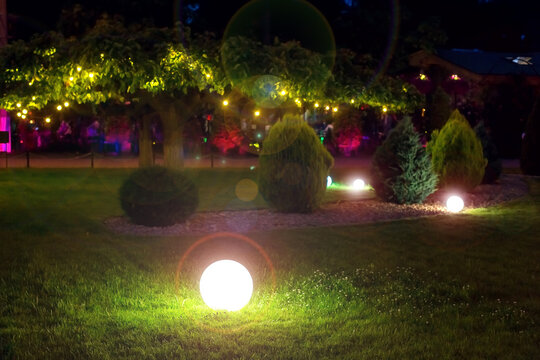 illumination park light glare  garden with electric ground ball lantern with stone mulch and thuja bushes in outdoor landscaped park with garland of warm light bulbs, illuminate evening scene nobody.
