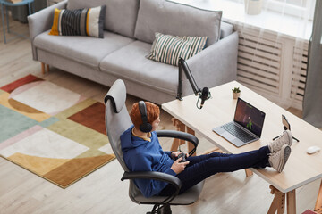 Wide angle above view at red haired teenage boy playing video games via laptop while relaxing in designer room, copy space © Seventyfour