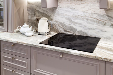 beautiful closeup of custom designed kitchen, with marble looking quartz countertop and backsplash. cream electric kettle with porcelain tea accessories on the marble countertop next to ceramic hob - obrazy, fototapety, plakaty