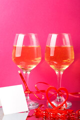 Two glasses with champagne tied with red ribbons on a pink background next to a postcard.