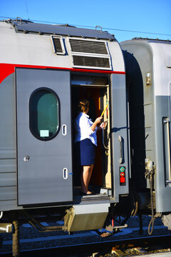 Railway cars. A female conductor stands in the doorway with a signal flag in her hand. Long-distance train, short stop at the railway station.
