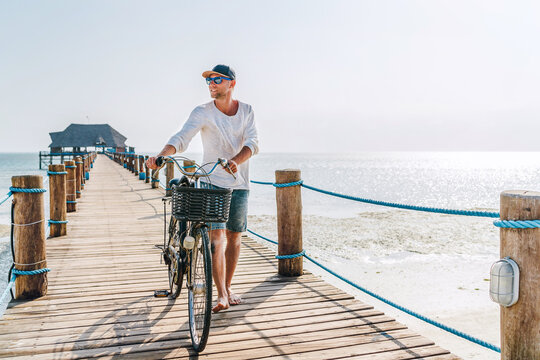 Portrait of a happy smiling barefoot man dressed in light summer clothes and sunglasses walking with a bicycle on the wooden sea pier. Careless vacation in tropical countries concept image.