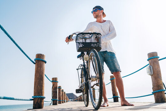Portrait of a happy smiling barefoot man dressed in light summer clothes and sunglasses riding a bicycle on the wooden sea pier. Careless vacation in tropical countries concept image.